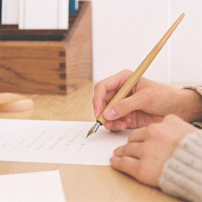 Person writing with wooden dip pen on paper on wooden table