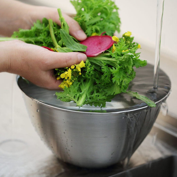 Person rinsing fresh greens over a metal colander on a light background