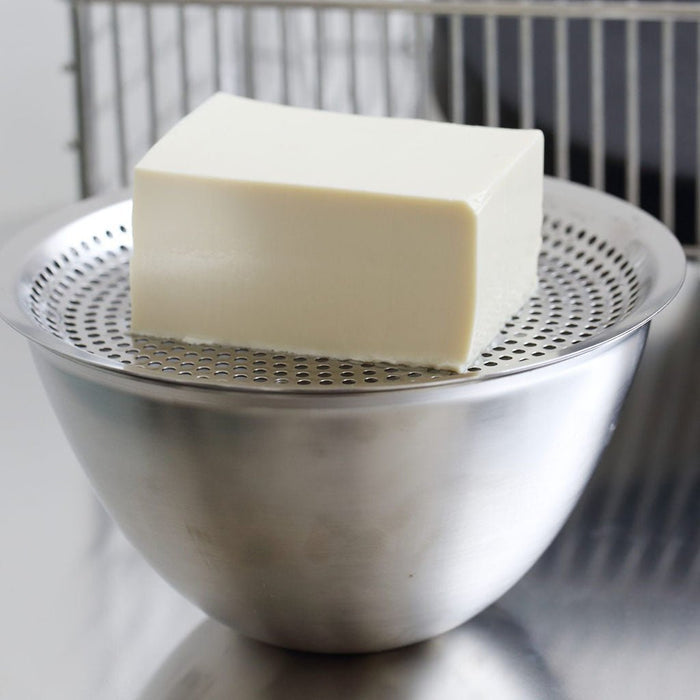 Block of tofu draining on flat strainer on top of steel bowl in kitchen