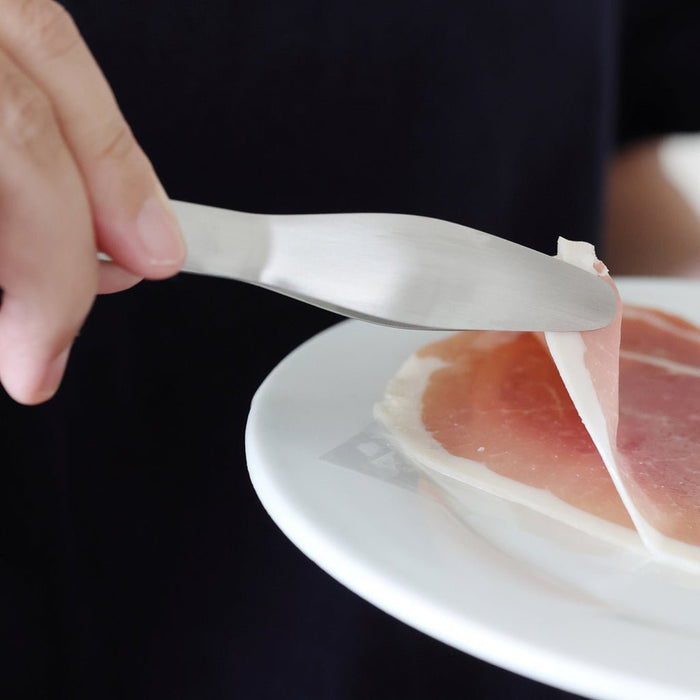 Person lifting a slice of prosciutto with a pair of tongs on a white plate.