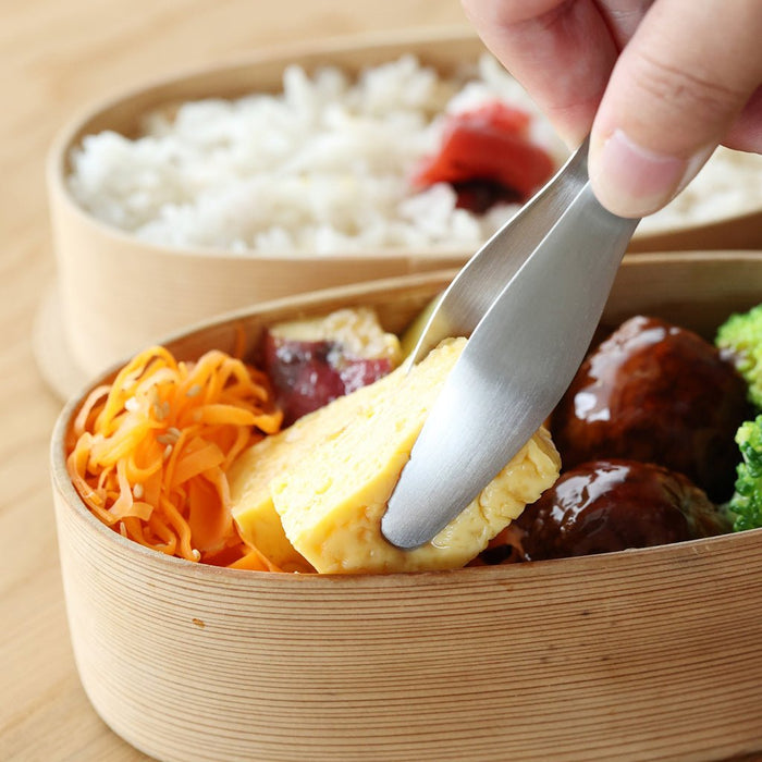 Bento box with food and a hand using tongs to pick up a piece of tofu.