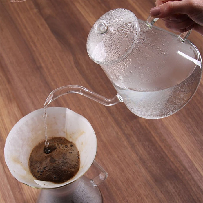 Clear glass teapot pouring water into a coffee filter with coffee grounds on a wooden surface.