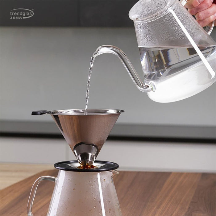 Clear glass kettle pouring water into a metal coffee filter on a wooden surface.